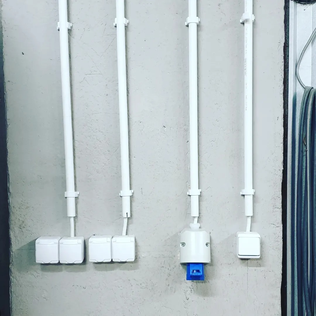 Białe rury instalacyjne przymocowane do szarej ściany, prowadzące do gniazdek i łączników elektrycznych, w tym jedno gniazdko z niebieskim wkładem.