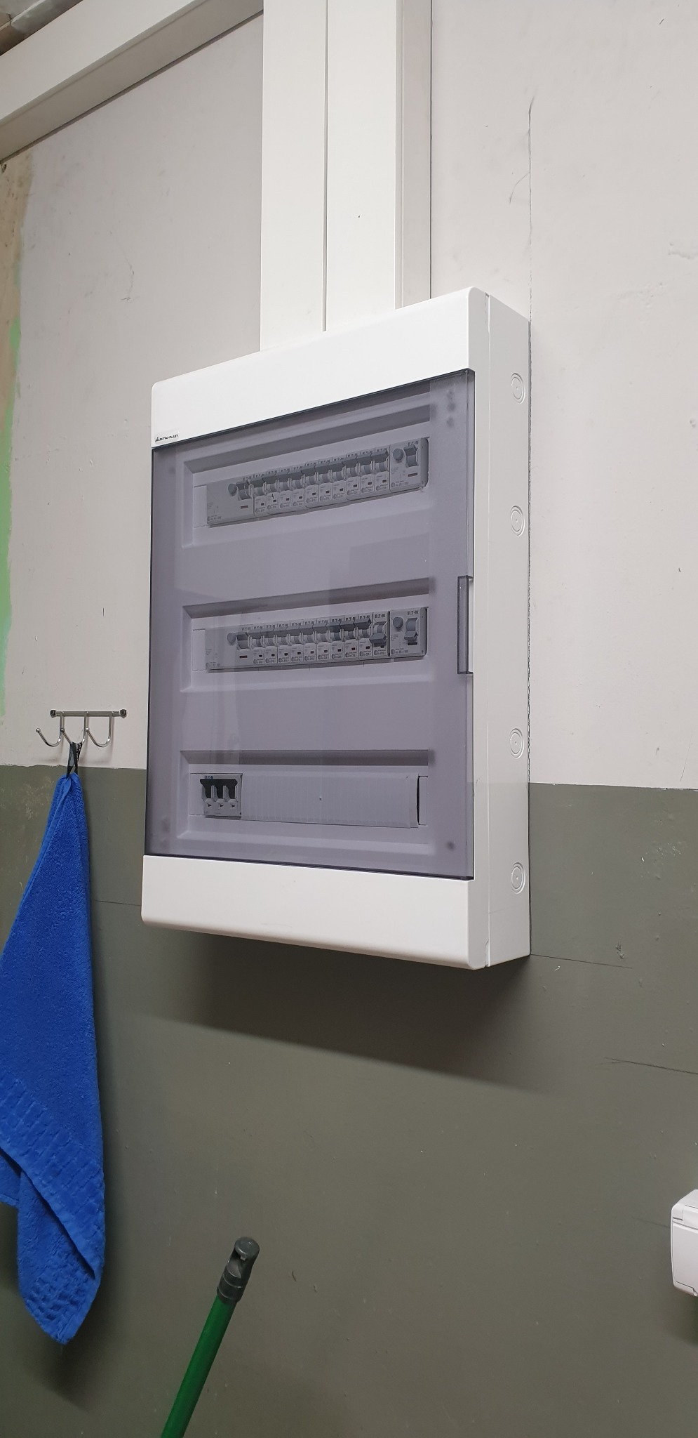 Zamknięta, biała skrzynka rozdzielcza z widocznymi bezpiecznikami, zamontowana na dwukolorowej ścianie, z niebieskim ręcznikiem wiszącym obok na metalowym wieszaku.