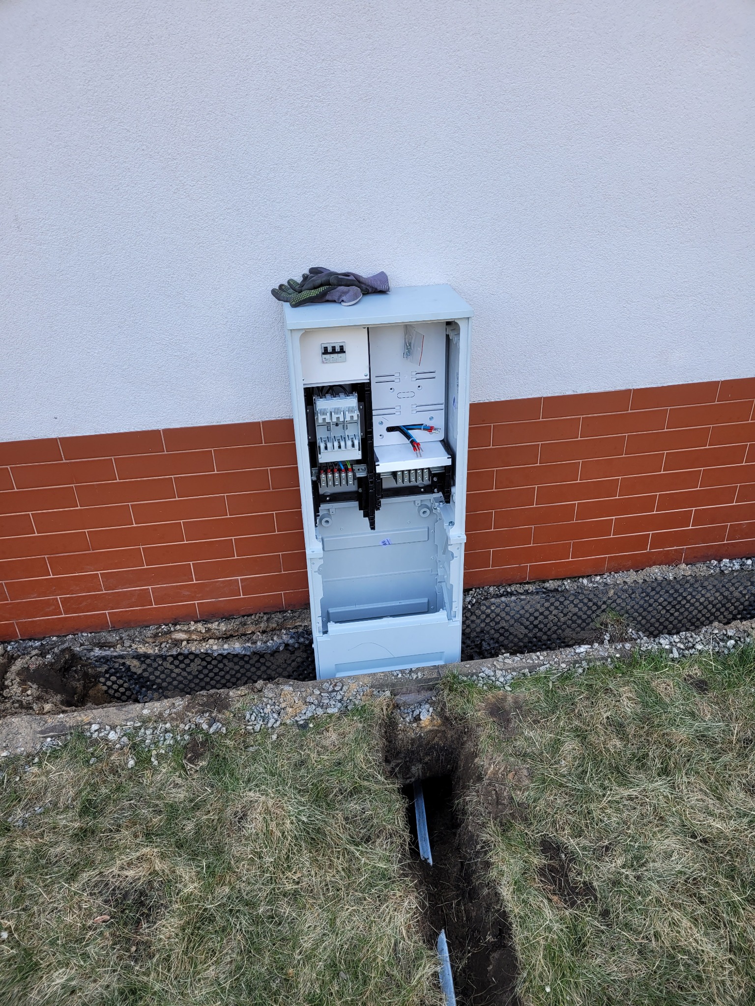 Otwarta skrzynka elektryczna, częściowo zamontowana na elewacji budynku, z widocznymi przewodami i elementami instalacji wewnątrz, obok wykopany rów z rurami, rękawice robocze leżące na górze...