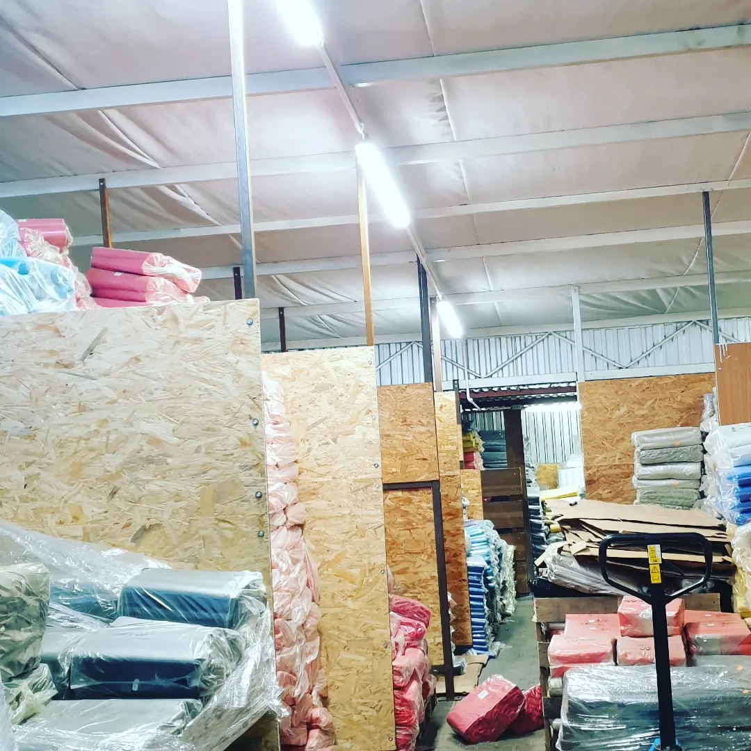 Wnętrze magazynu z paletami wypełnionymi towarem, widoczne oświetlenie sufitowe i wózek paletowy.