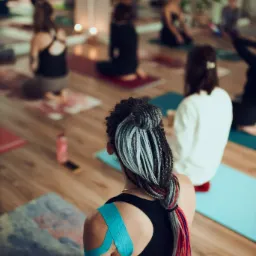 Grupa osób ćwiczy w sali, widok zza pleców kobiety siedzącej na macie z plastrami kinesiotaping na ramieniu i kolorowymi warkoczykami.