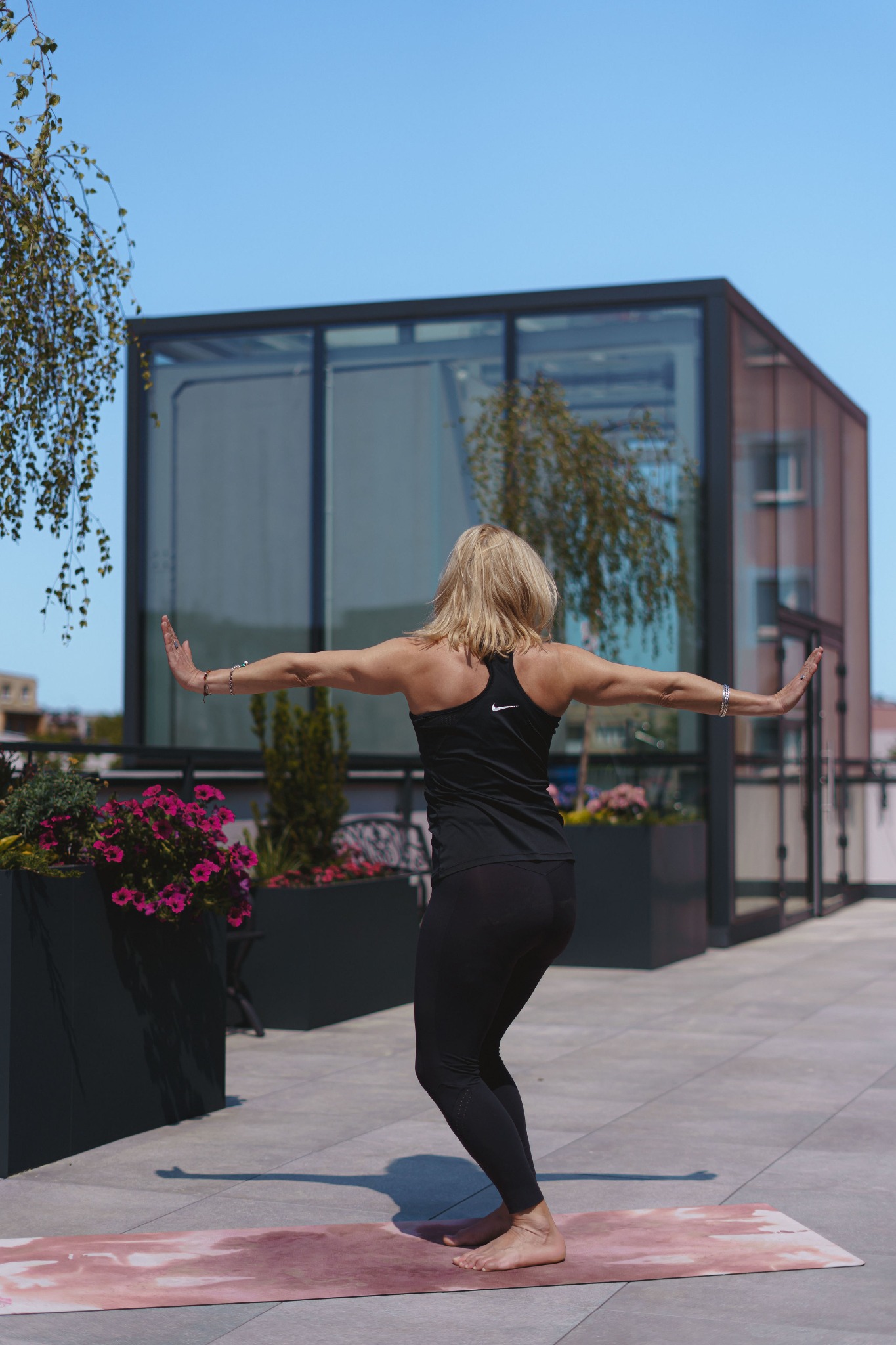 Kobieta w czarnym stroju sportowym wykonuje ćwiczenie na macie na tarasie z widokiem na nowoczesny budynek z przeszkloną fasadą i roślinami w donicach.