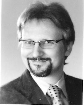 Portret biznesowy w sepii: mężczyzna w okularach, z brodą i wąsami, w garniturze i krawacie w kropki, uśmiechnięty, z delikatnym cieniowaniem na twarzy.