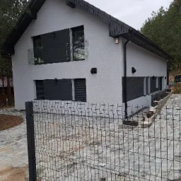 Dwukondygnacyjny dom z szarą elewacją, czarnymi akcentami i szklanymi balustradami, widziany zza metalowego ogrodzenia panelowego na tle drzew.