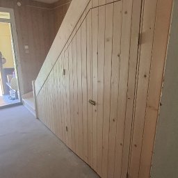 INOMHUS Prace Wykończeniowe i Budownictwo - Drewniane schody z zabudową szafy pod schodami, wykończone boazerią. Widoczne drzwi wejściowe i surowy strop. Wykończenie wnętrza w naturalnym stylu.