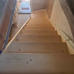 INOMHUS Prace Wykończeniowe i Budownictwo - Drewniane schody strychowe prowadzące na poddasze, widok z dołu. Surowe drewno, widoczne łączenia. W tle okno dachowe i ocieplone ściany poddasza.