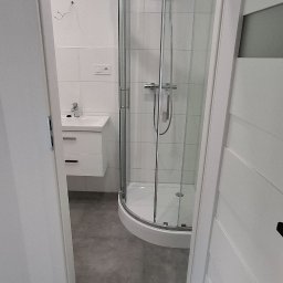 INOMHUS Prace Wykończeniowe i Budownictwo - Nowoczesna łazienka z kabiną prysznicową narożną, białą umywalką z szafką i szarą podłogą, widziana zza uchylonych białych drzwi.