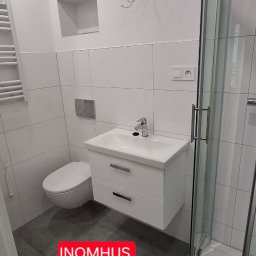 INOMHUS Prace Wykończeniowe i Budownictwo - Nowoczesna łazienka z białą armaturą: wisząca szafka z umywalką, toaleta i kabina prysznicowa. Szare płytki podłogowe i białe płytki ścienne.