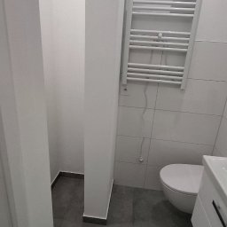 INOMHUS Prace Wykończeniowe i Budownictwo - Nowoczesna, minimalistyczna łazienka w odcieniach szarości i bieli. Widoczna toaleta, grzejnik drabinkowy i umywalka z szafką. Szare płytki podłogowe i białe ściany.