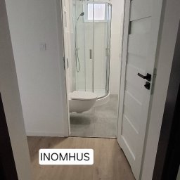 INOMHUS Prace Wykończeniowe i Budownictwo - Jasna łazienka po remoncie: kabina prysznicowa z hydromasażem, biała toaleta, drewnopodobna podłoga i białe drzwi z czarną klamką. Widok z korytarza.