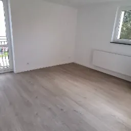 Jasne panele podłogowe w nowym, pustym pokoju z białymi ścianami, oknem i balkonem. Widoczne listwy przypodłogowe i grzejnik. Minimalistyczne wnętrze.