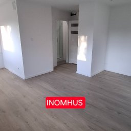 INOMHUS Prace Wykończeniowe i Budownictwo - Jasne panele podłogowe w pustym, odnowionym pokoju z białymi ścianami, widok na łazienkę z kabiną prysznicową. Minimalistyczny design wnętrza.