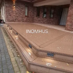 Nowoczesny taras kompozytowy z oświetleniem schodkowym w Debrznie, przylegający do ceglanego domu z ciemną dachówką. Widok z poziomu kostki brukowej.