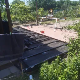 Montaż tarasu kompozytowego na podłożu z czarnej membrany, piła ukośnica na stojaku w tle, widok na ogród.