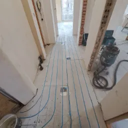 Montaż ogrzewania podłogowego w korytarzu, widoczne niebieskie rurki układane na płytach izolacyjnych, w tle odkurzacz przemysłowy i narzędzia, surowy stan wykończenia.