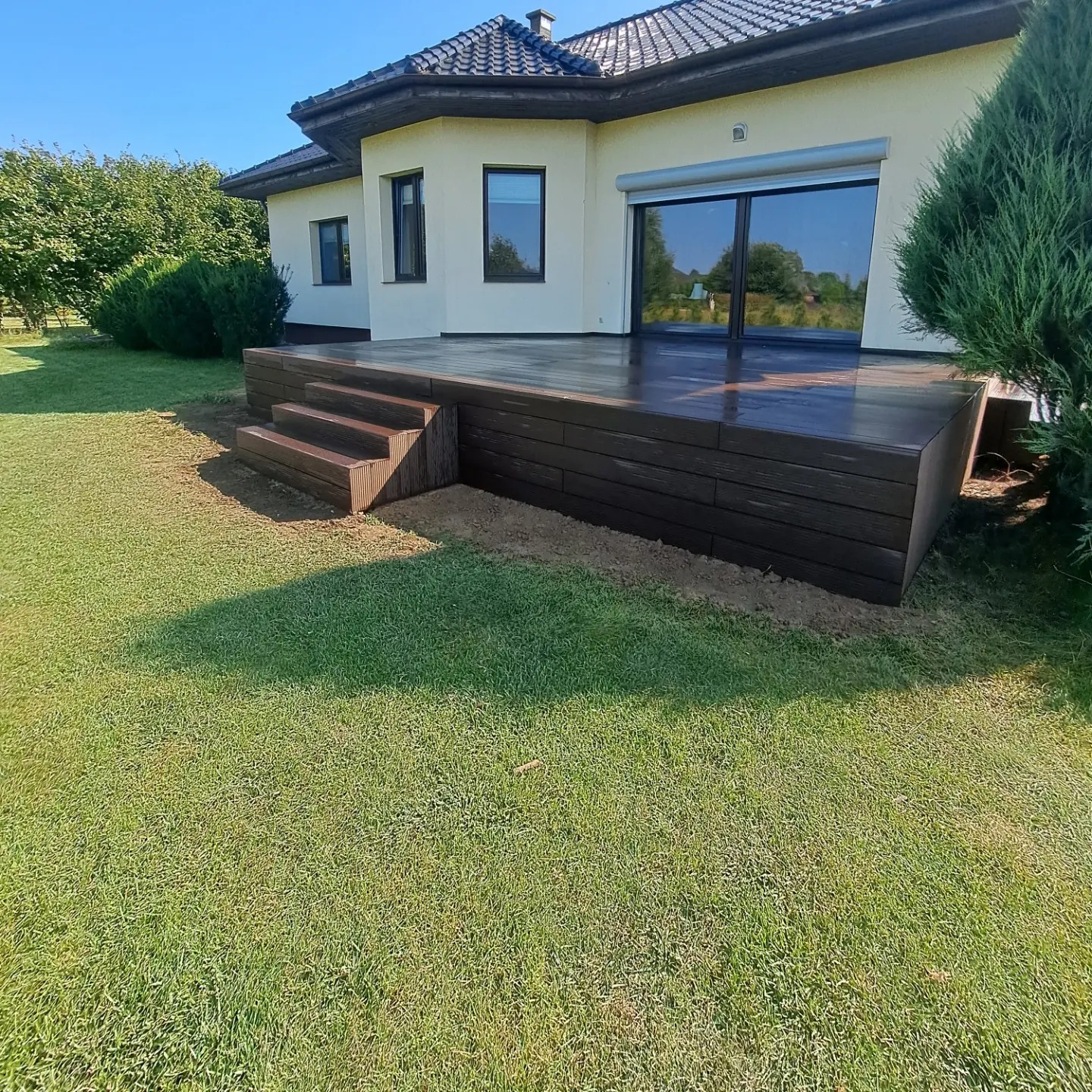 Nowoczesny taras z kompozytu w kolorze ciemnego drewna, z niskimi schodami, przylegający do jasnego domu z dużymi oknami przesuwnymi i zielonym trawnikiem.
