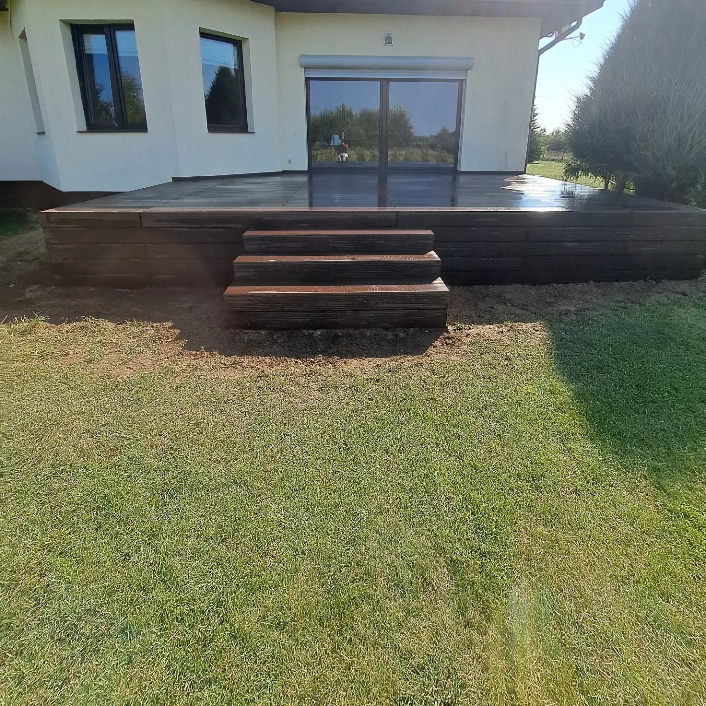 Nowoczesny taras z kompozytowych desek w kolorze ciemnego drewna, z trzema stopniami prowadzącymi do drzwi tarasowych, otoczony zielonym trawnikiem i fragmentem białego domu z dużymi oknami.