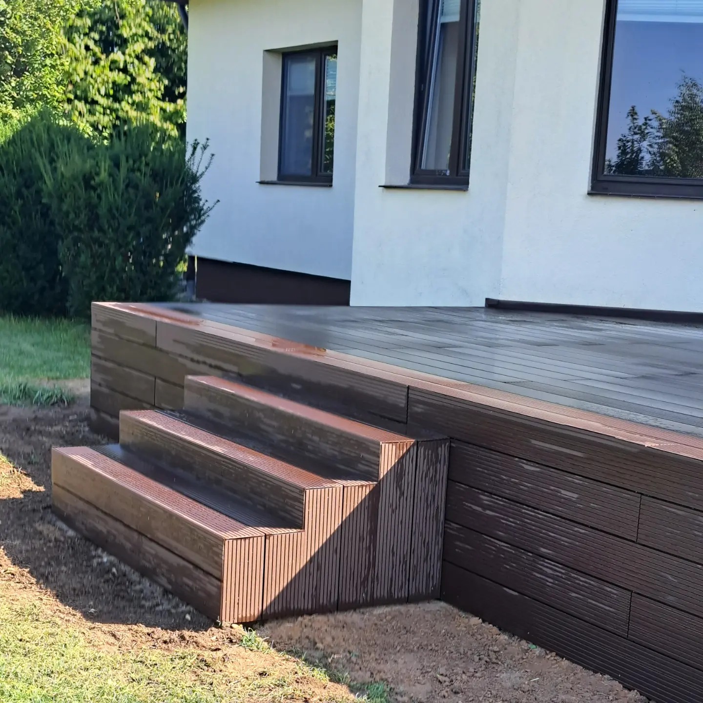 Nowoczesny taras z kompozytowych desek w kolorze ciemnego drewna, z trzema schodkami prowadzącymi do domu z białym tynkiem, widok z boku, trawa i krzewy w tle.
