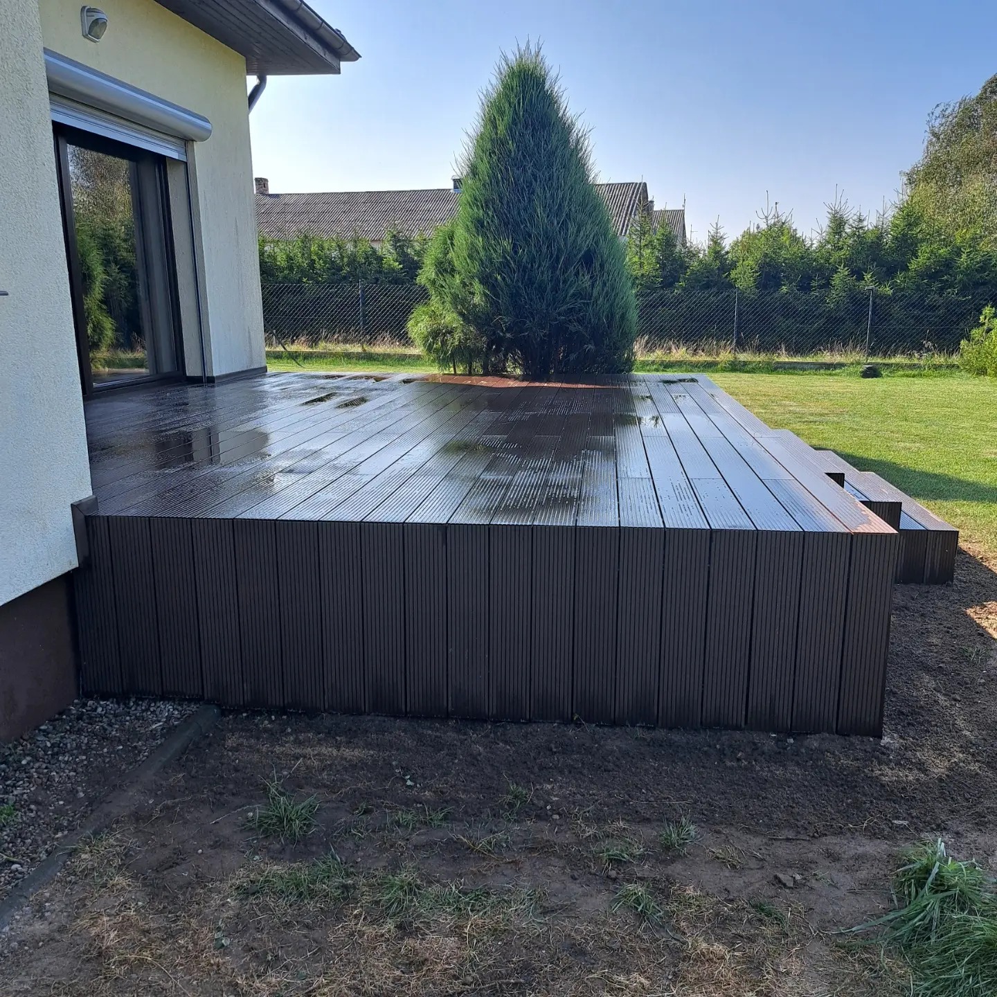 Nowo wybudowany, ciemnobrązowy taras kompozytowy przy domu jednorodzinnym, z widocznymi śladami wilgoci na deskach, otoczony świeżo przekopaną ziemią i trawnikiem w słoneczny dzień.