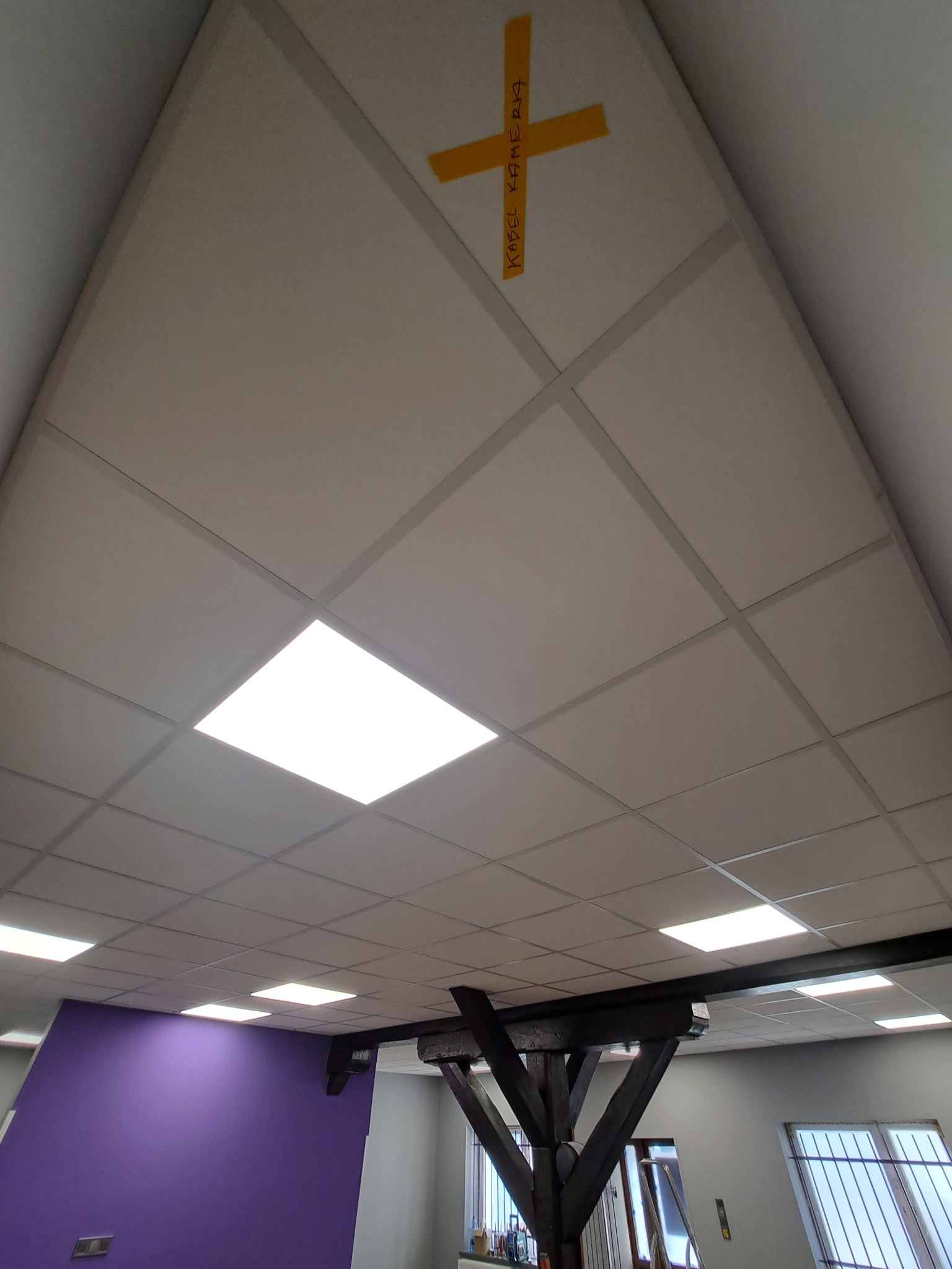 Sufit podwieszany kasetonowy z kwadratowymi panelami i wbudowanym oświetleniem LED, na jednym z paneli żółta taśma z napisem 'KABEL KAMERA', widoczne elementy konstrukcyjne podtrzymujące sufit...