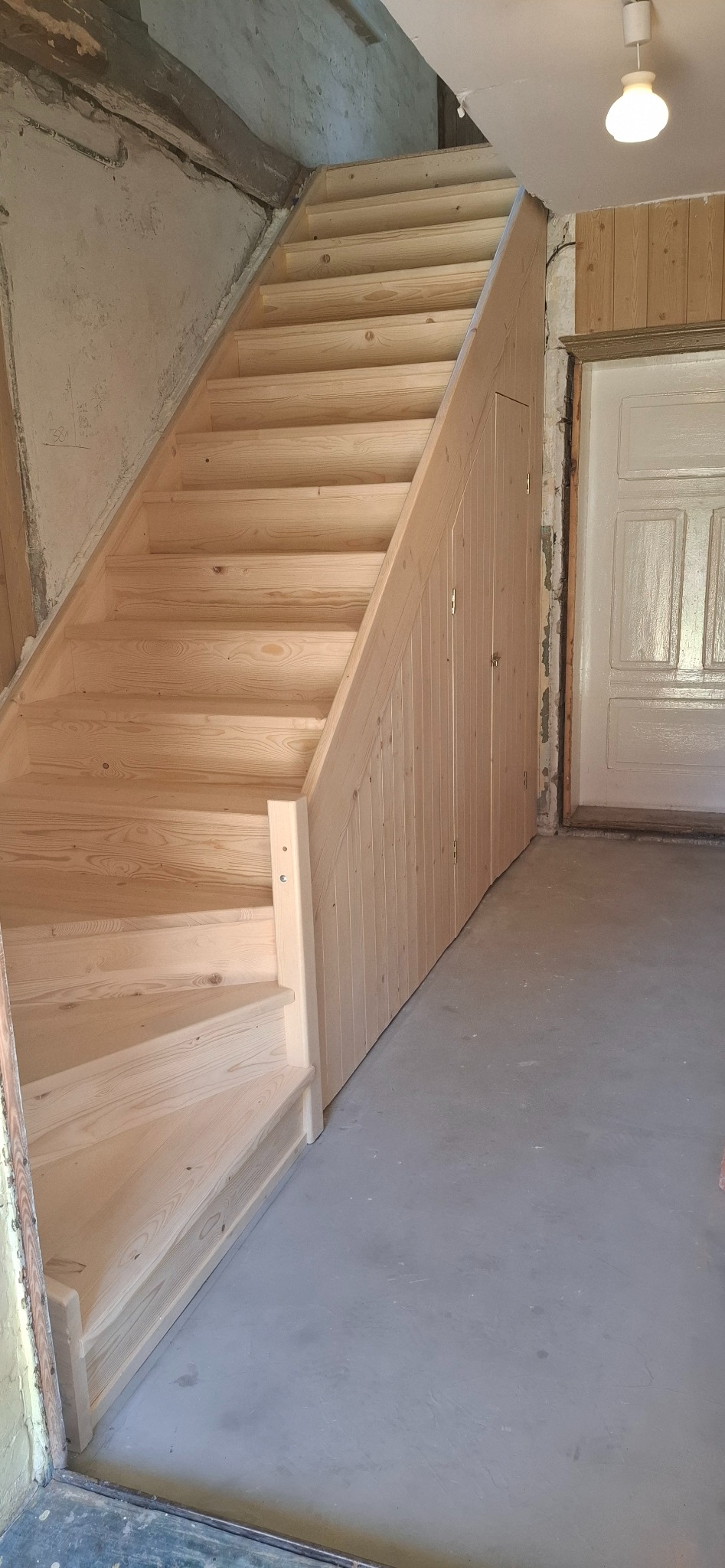 Jasne, drewniane schody ze schowkiem pod biegiem, widoczne na tle surowych ścian i fragmentu drzwi. Naturalne światło podkreśla strukturę drewna.