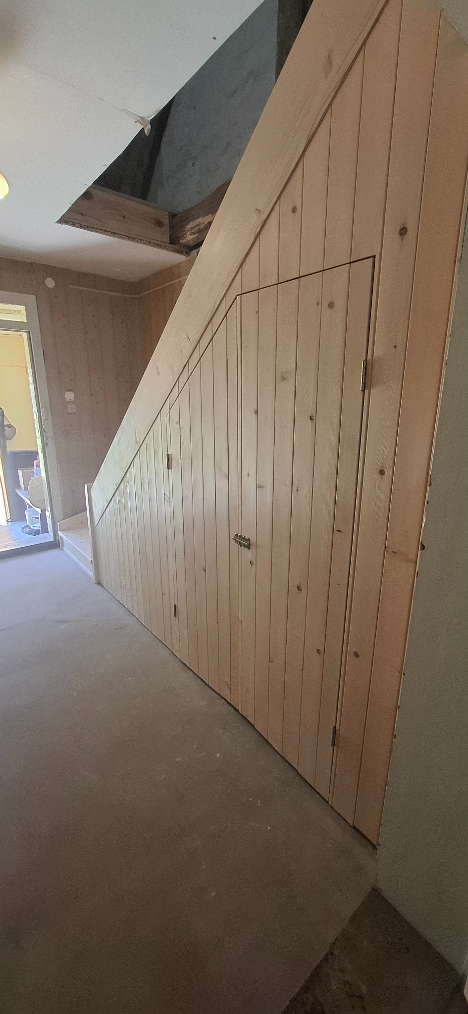 Drewniane schody z zabudową szafy pod schodami, wykończone boazerią. Widoczne drzwi wejściowe i surowy strop. Wykończenie wnętrza w naturalnym stylu.