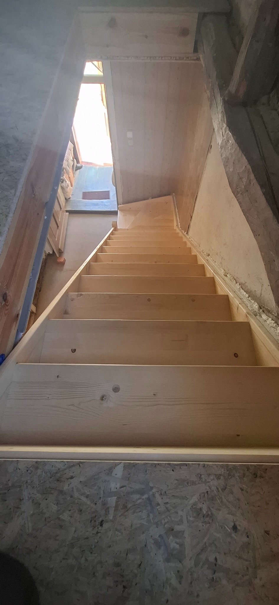 Drewniane schody strychowe prowadzące na poddasze, widok z dołu. Surowe drewno, widoczne łączenia. W tle okno dachowe i ocieplone ściany poddasza.