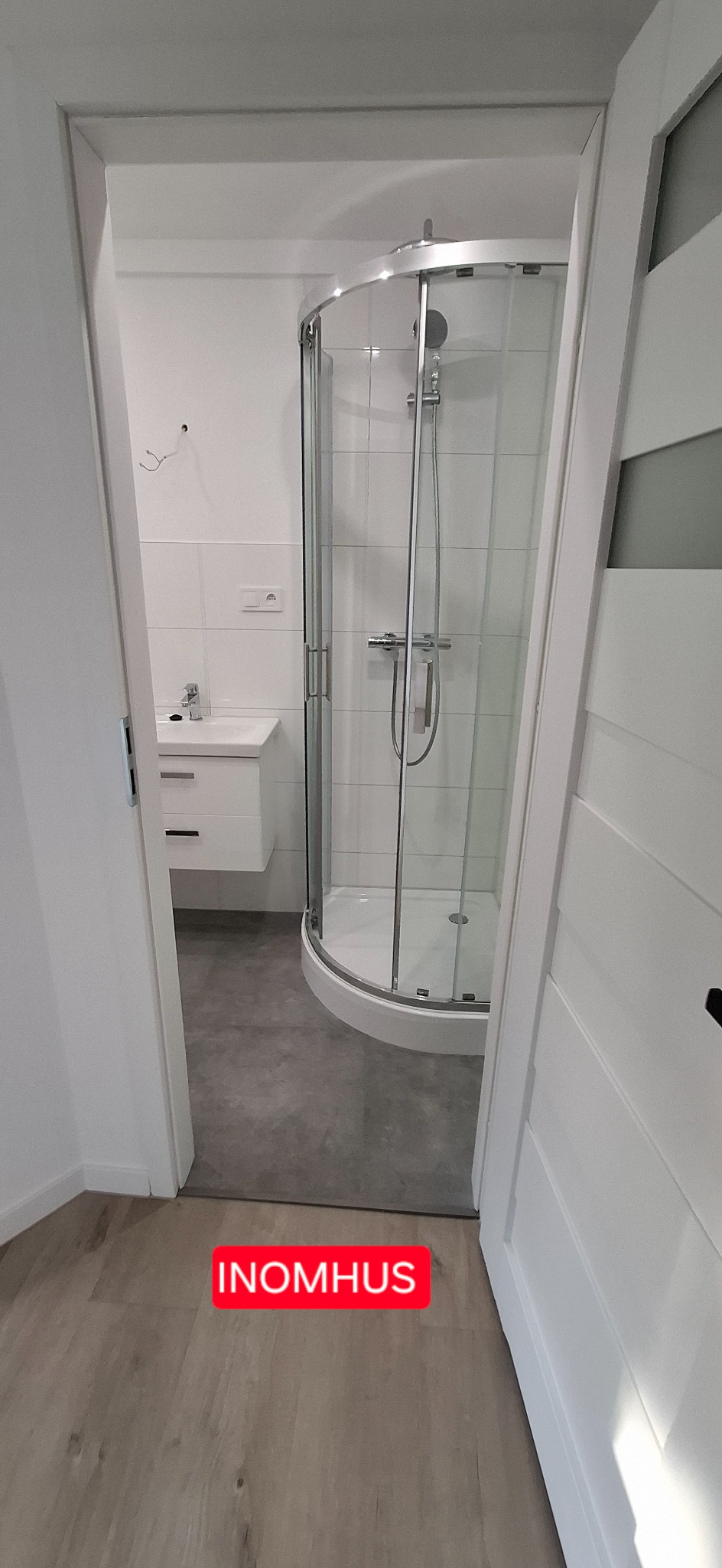 Nowoczesna łazienka z kabiną prysznicową narożną, białą umywalką z szafką i szarą podłogą, widziana zza uchylonych białych drzwi.