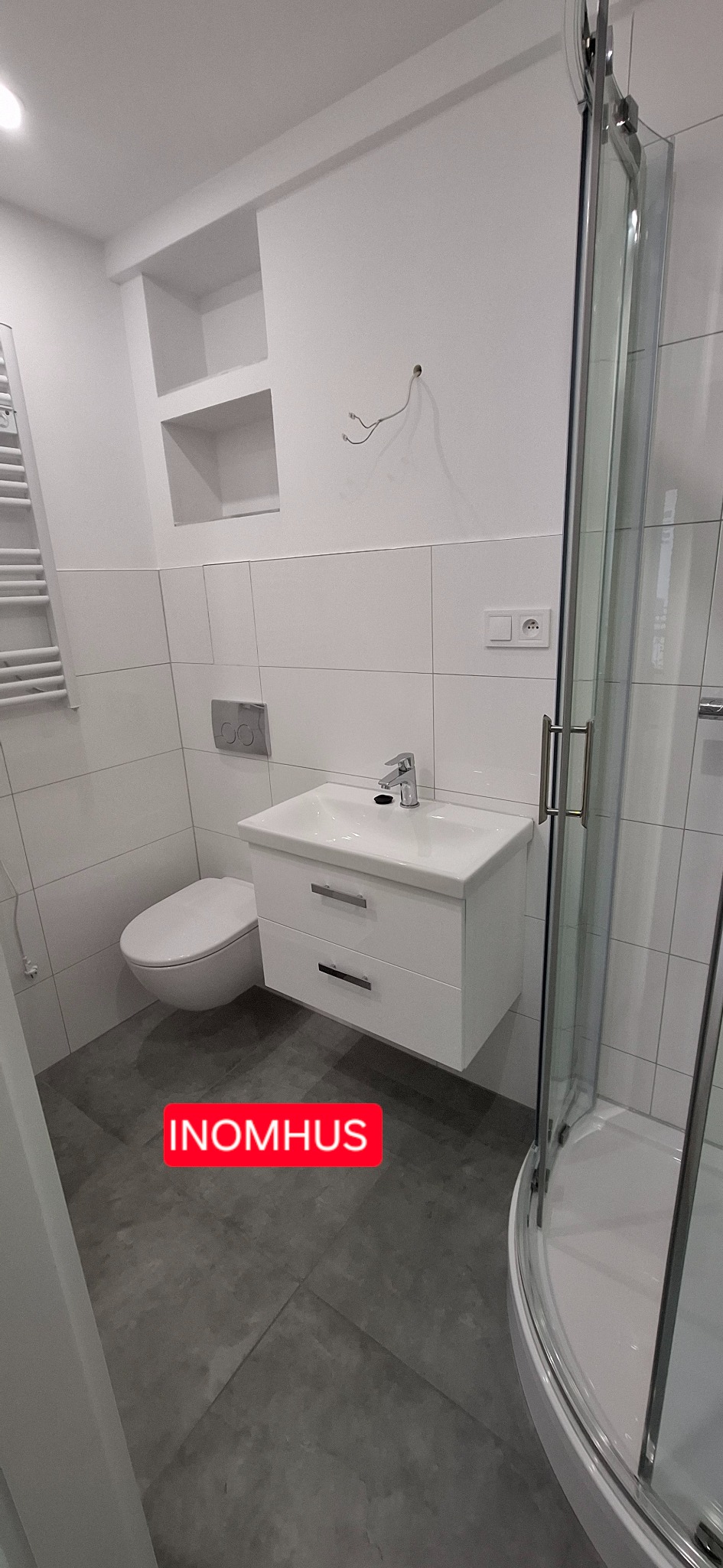 Nowoczesna łazienka z białą armaturą: wisząca szafka z umywalką, toaleta i kabina prysznicowa. Szare płytki podłogowe i białe płytki ścienne.