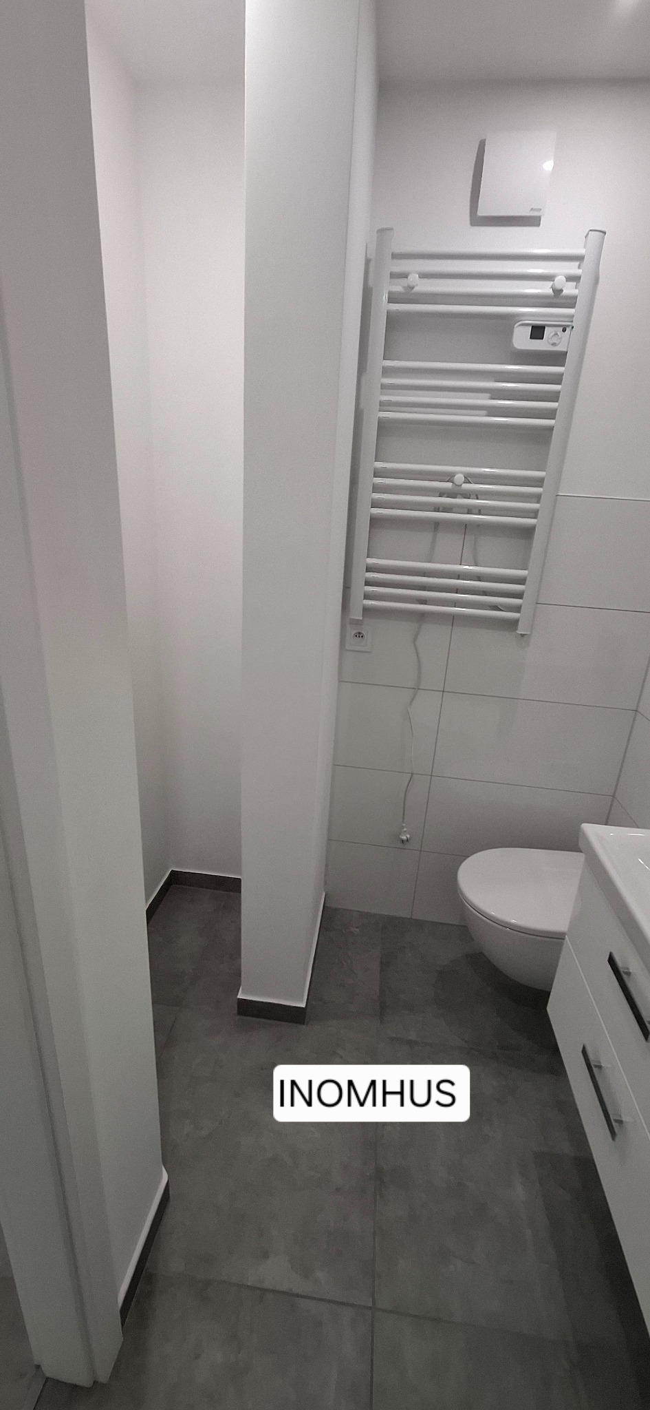 Nowoczesna, minimalistyczna łazienka w odcieniach szarości i bieli. Widoczna toaleta, grzejnik drabinkowy i umywalka z szafką. Szare płytki podłogowe i białe ściany.