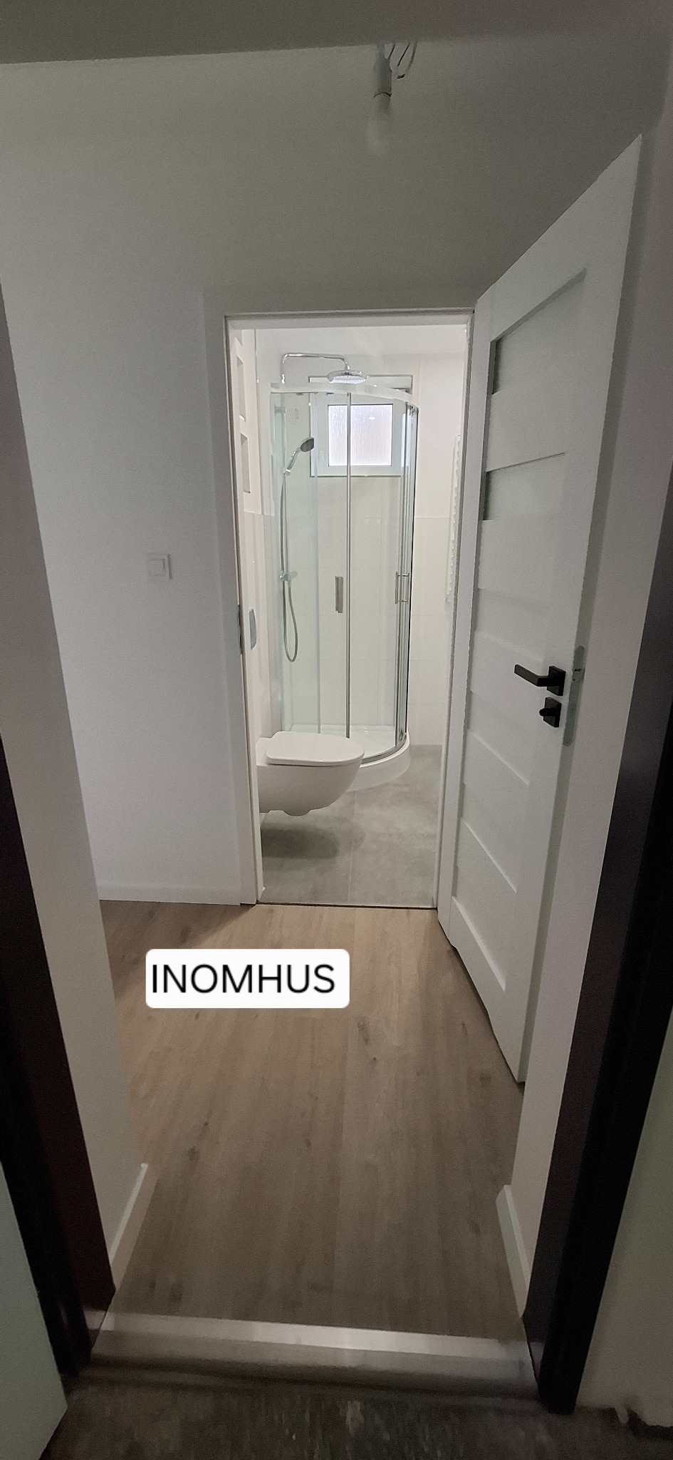 Jasna łazienka po remoncie: kabina prysznicowa z hydromasażem, biała toaleta, drewnopodobna podłoga i białe drzwi z czarną klamką. Widok z korytarza.