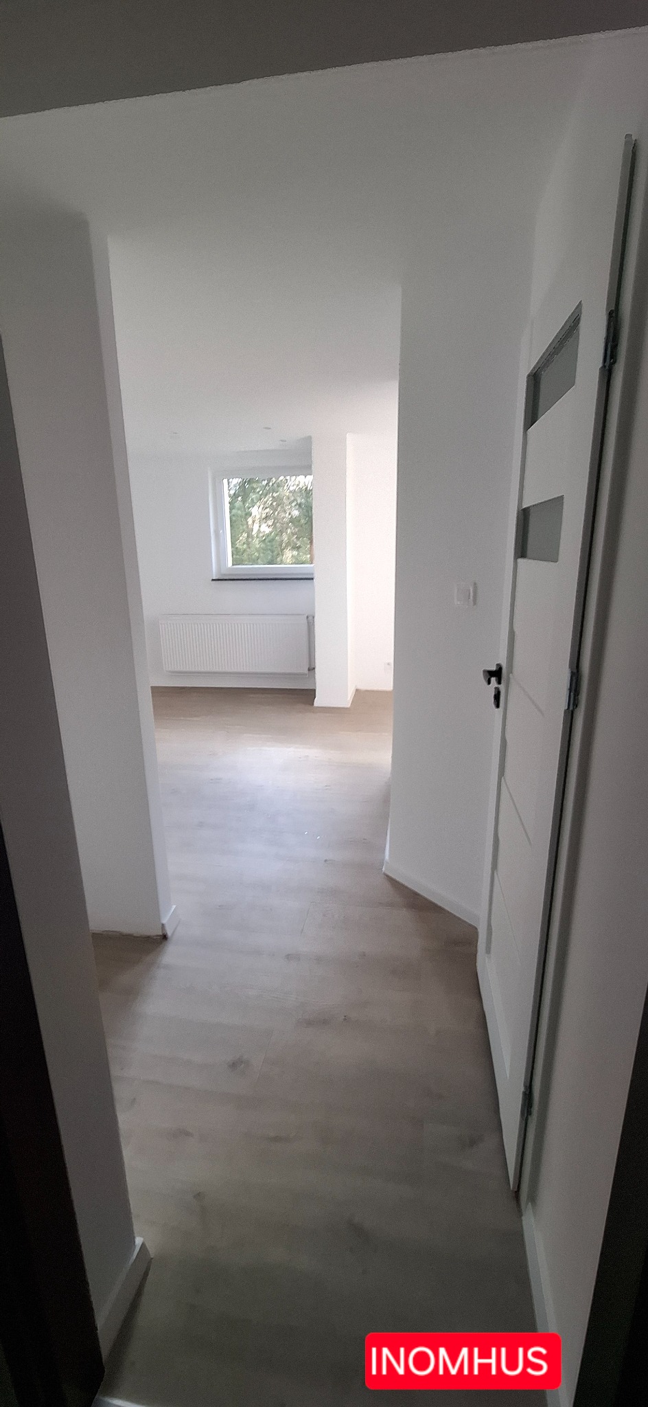 Jasne wnętrze po remoncie: świeżo pomalowane ściany, panele podłogowe w odcieniach beżu, nowe drzwi z przeszkleniami i widok na okno z grzejnikiem.