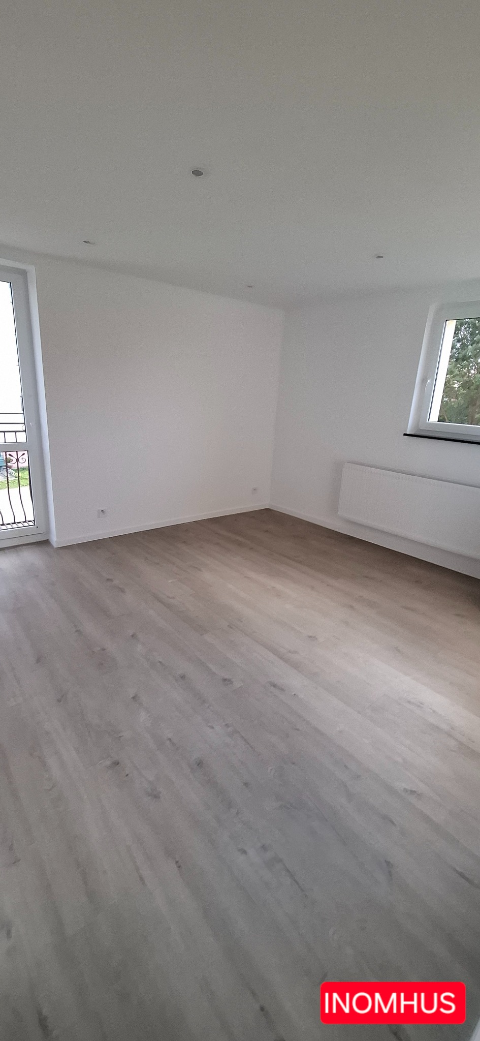 Jasne panele podłogowe w nowym, pustym pokoju z białymi ścianami, oknem i balkonem. Widoczne listwy przypodłogowe i grzejnik. Minimalistyczne wnętrze.