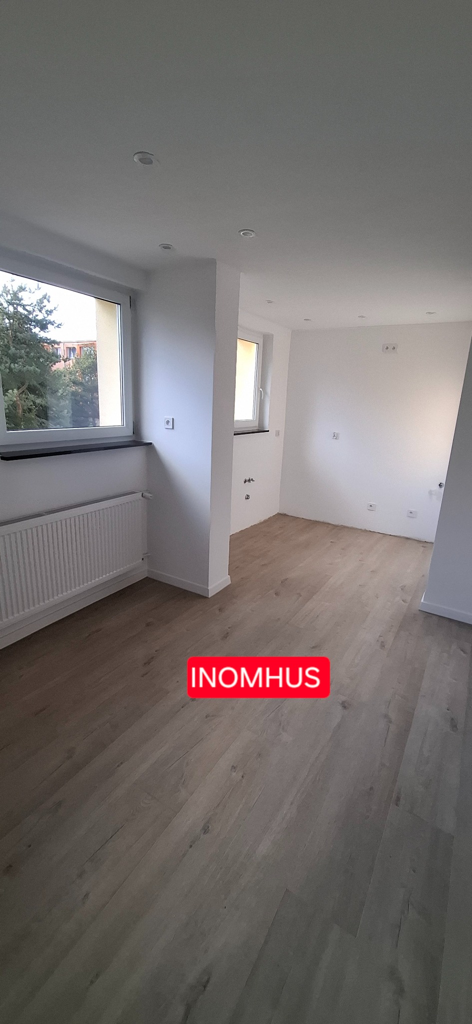 Jasne panele podłogowe w nowym wnętrzu w Debrznie. Świeżo wykończone ściany, okna i grzejnik. Widok na dwa pomieszczenia i instalacje elektryczne.