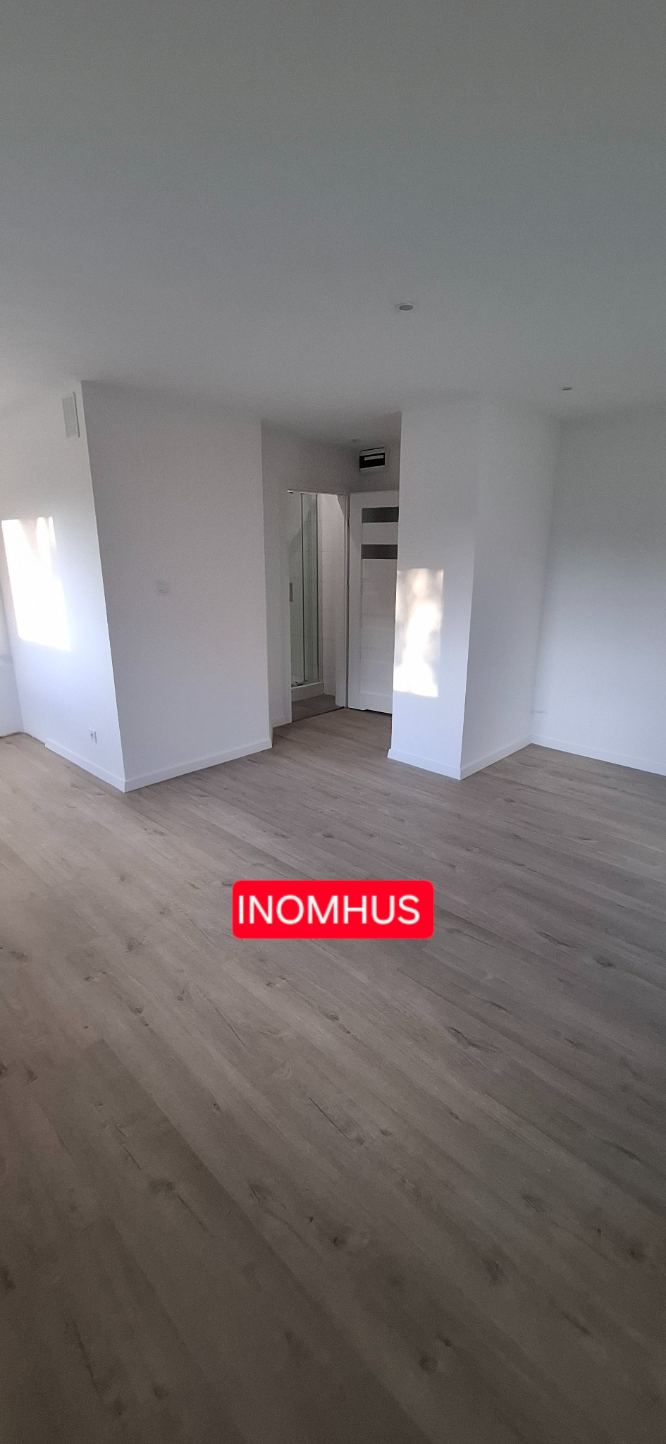 Jasne panele podłogowe w pustym, odnowionym pokoju z białymi ścianami, widok na łazienkę z kabiną prysznicową. Minimalistyczny design wnętrza.