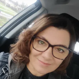 Autoportret kobiety w okularach w samochodzie, z widokiem na zewnątrz przez okno. Ubrana w skórzaną kurtkę. Uśmiechnięta, z bliska.