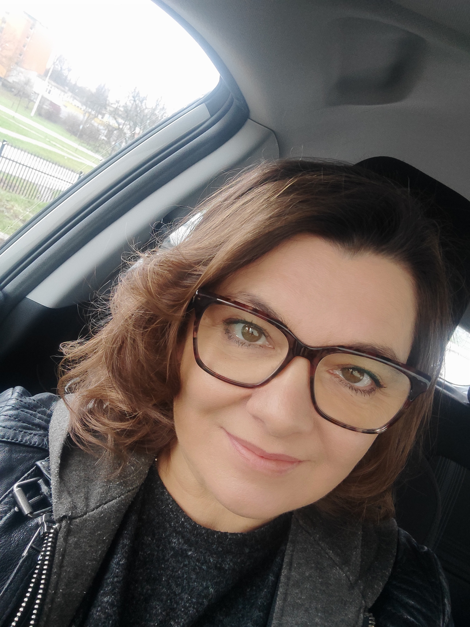 Autoportret kobiety w okularach w samochodzie, z widokiem na zewnątrz przez okno. Ubrana w skórzaną kurtkę. Uśmiechnięta, z bliska.