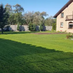 Rozległy, równo przystrzyżony trawnik przed domem z cegły, w tle betonowe ogrodzenie i drzewa, widoczny świeżo przygotowany fragment ziemi pod nasadzenia.