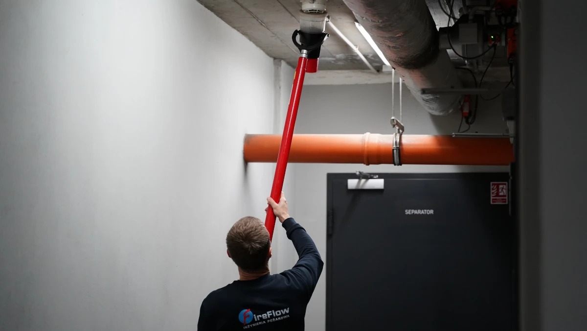 Czyszczenie czujnika dymu na suficie za pomocą specjalistycznego narzędzia z długą czerwoną rurą. Widoczny pracownik w uniformie firmy FireFlow.