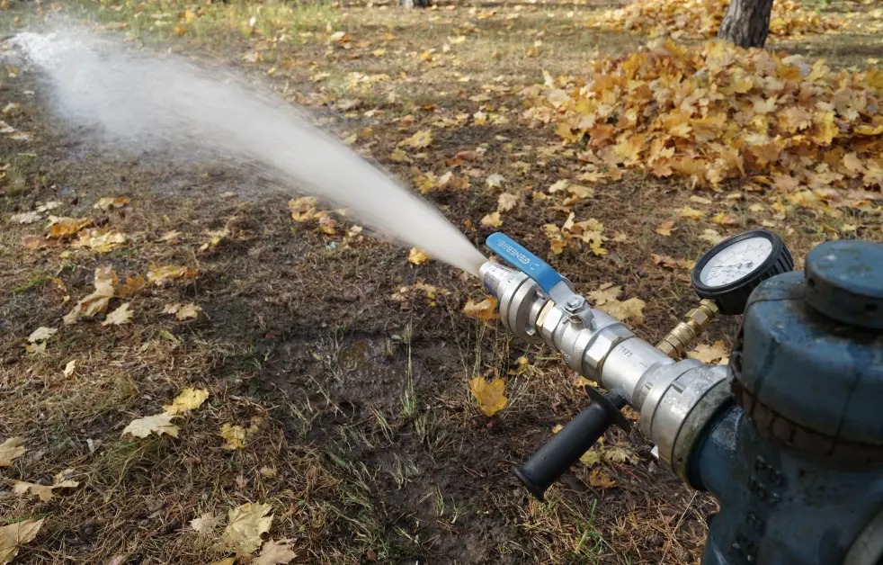 Hydrant zewnętrzny w akcji, woda pod ciśnieniem tryska na tle jesiennych liści. Manometr wskazuje ciśnienie. Element systemu ppoż. na zewnątrz budynku.