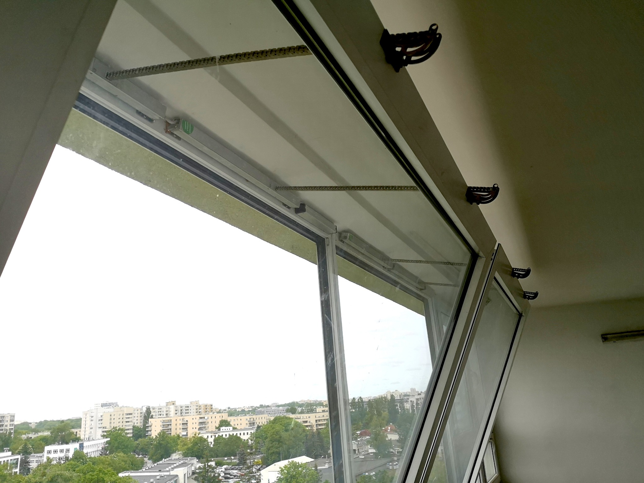 Otwarty uchylnie okno z widokiem na miasto. Widoczne elementy mechanizmu okiennego i uchwyty na suficie. Ujęcie z dołu, jasne światło dzienne.