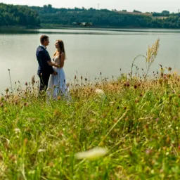 Para młoda w eleganckich strojach stoi na tle jeziora i zieleni, z dronem widocznym w tle, ujęcie z dołu przez wysokie trawy.
