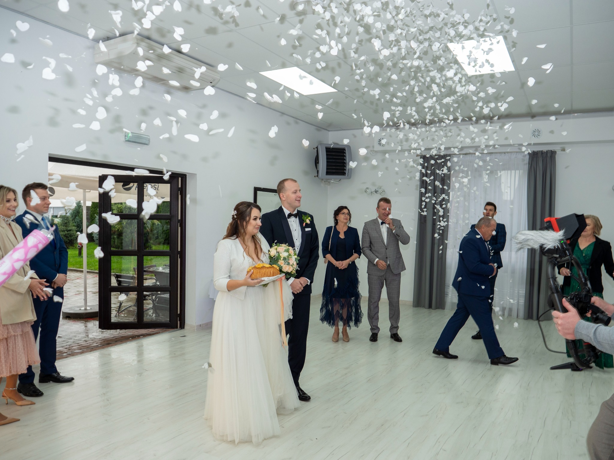 Para młoda w trakcie ceremonii powitalnej z chlebem i solą, obsypywana białymi płatkami, z gośćmi i kamerzystą w tle, jasne wnętrze sali.