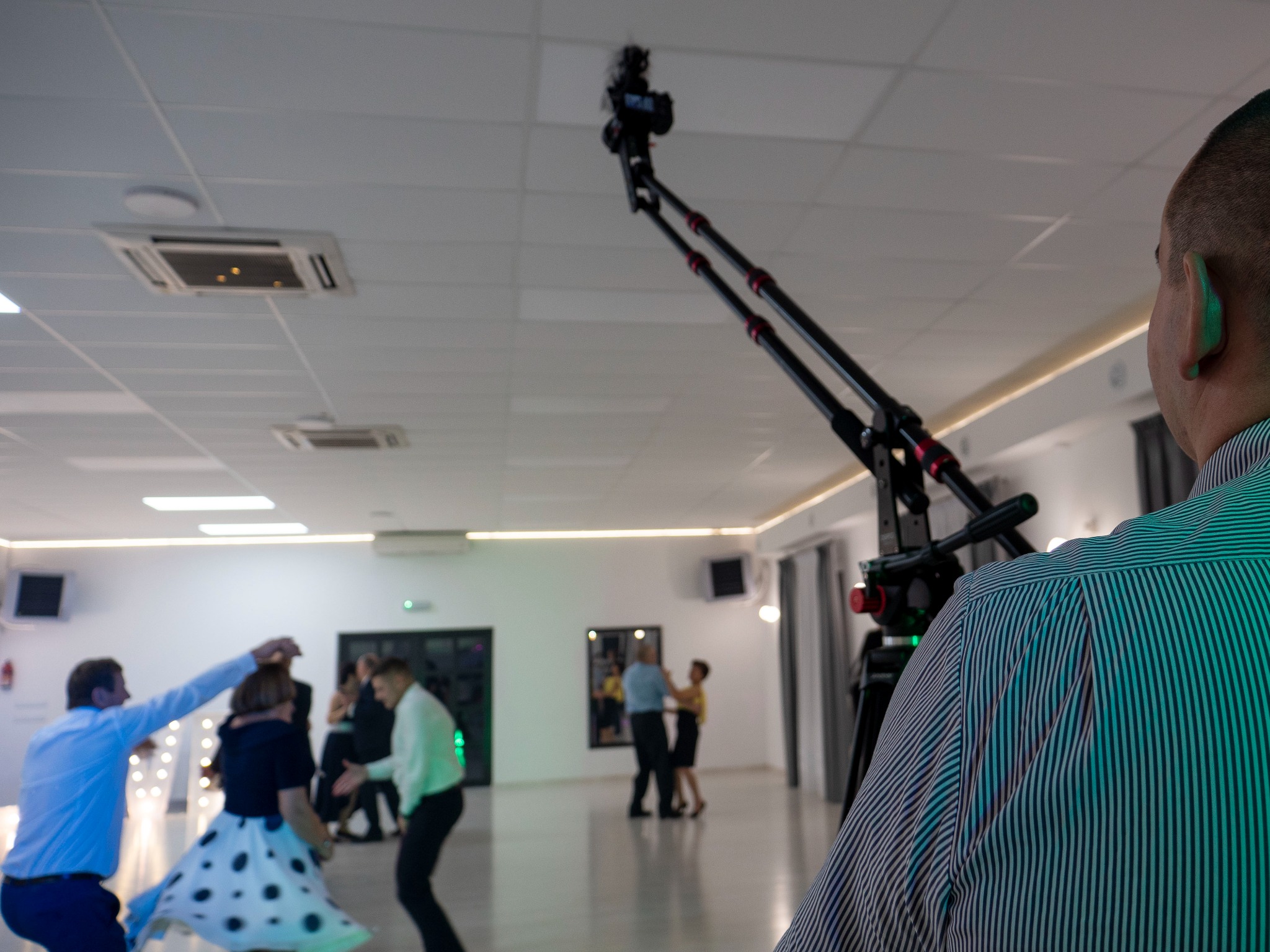 Kamera na wysięgniku nagrywa tańczących gości podczas imprezy, widok zza ramienia operatora w koszuli w paski.