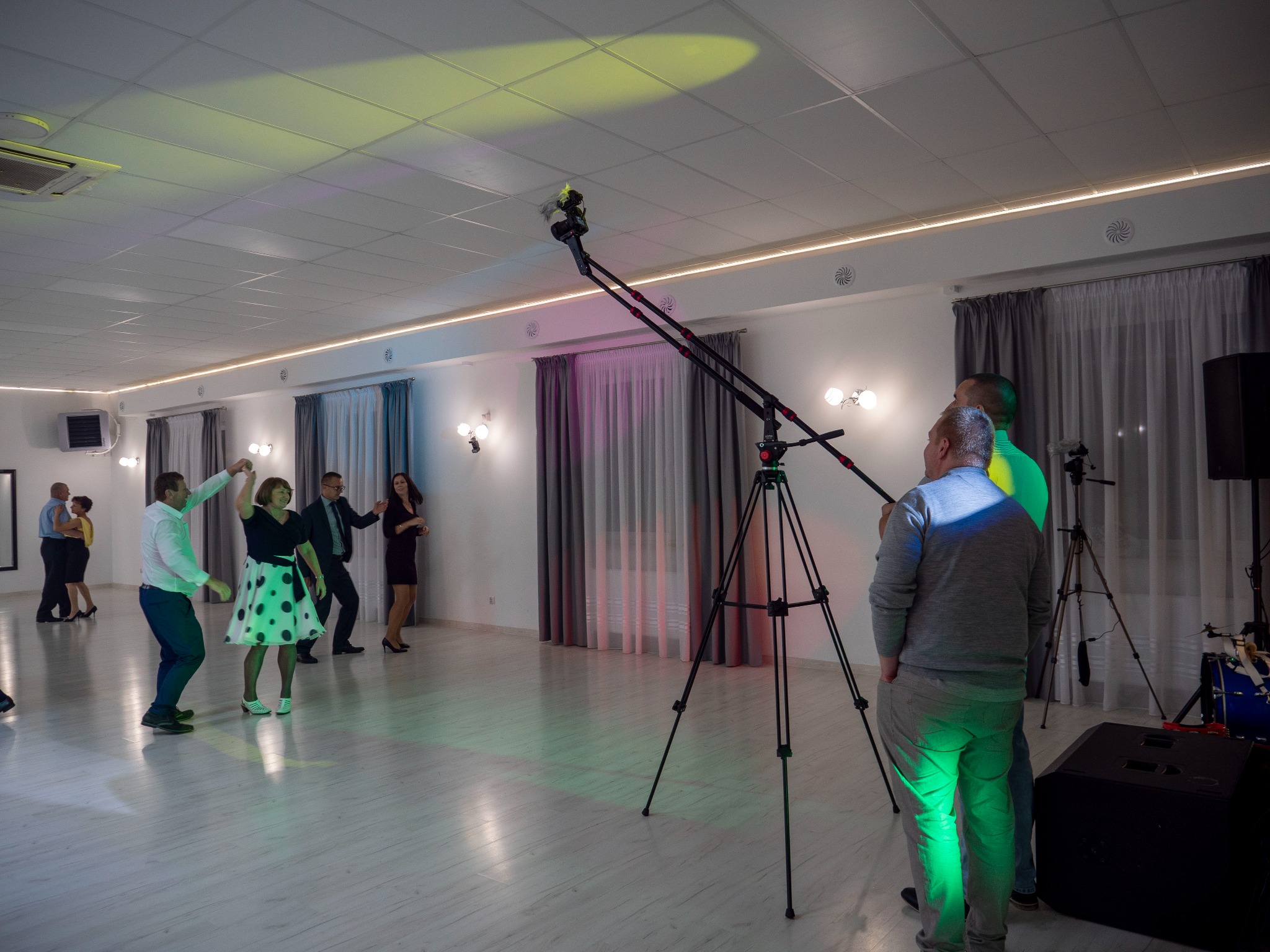 Sala balowa z tańczącymi osobami, oświetlona kolorowymi światłami, filmowana za pomocą kamery na wysięgniku. Widoczny operator kamery i sprzęt nagłośnieniowy.