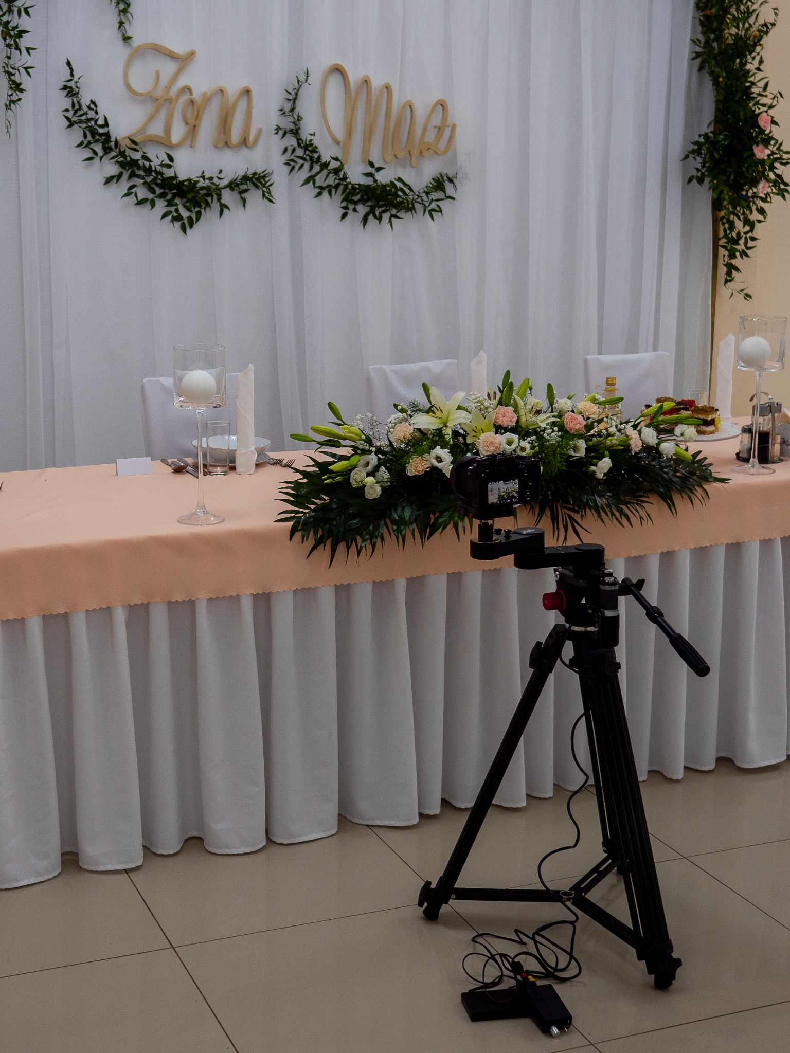 Kamera na statywie ustawiona przed udekorowanym stołem nowożeńców z napisem 'Żona Mąż', kwiatami, świecami i zastawą, gotowa do nagrywania.