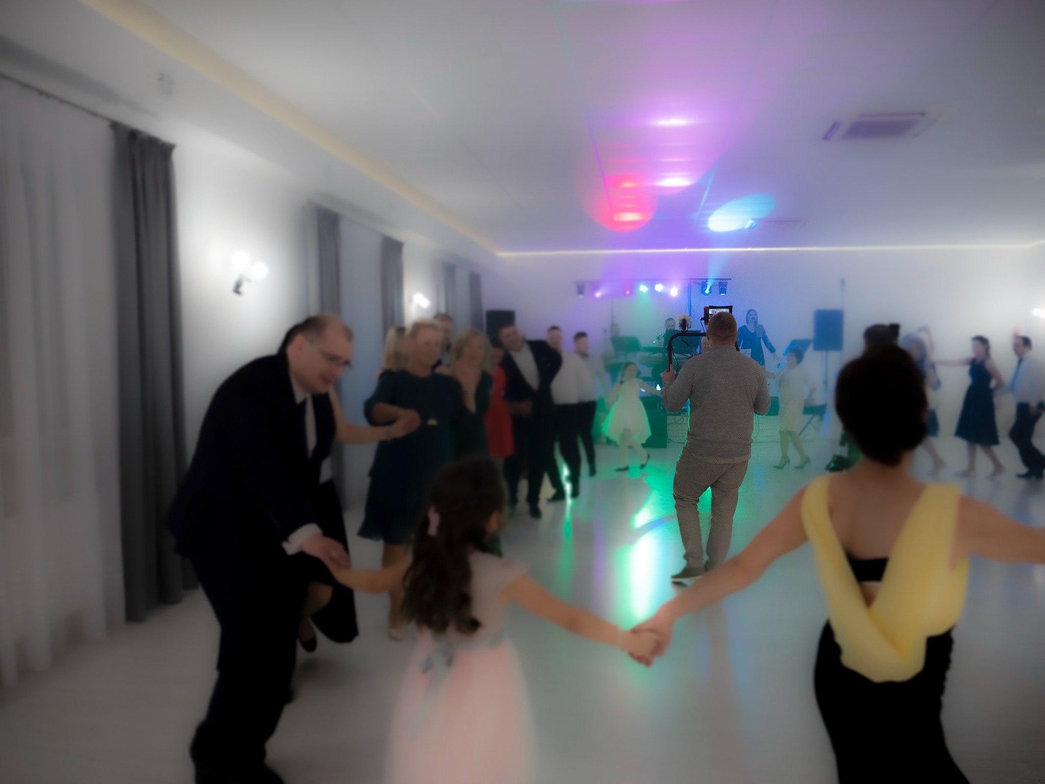 Goście weselni tańczą w sali oświetlonej kolorowymi światłami, w tle widoczny DJ z sprzętem.