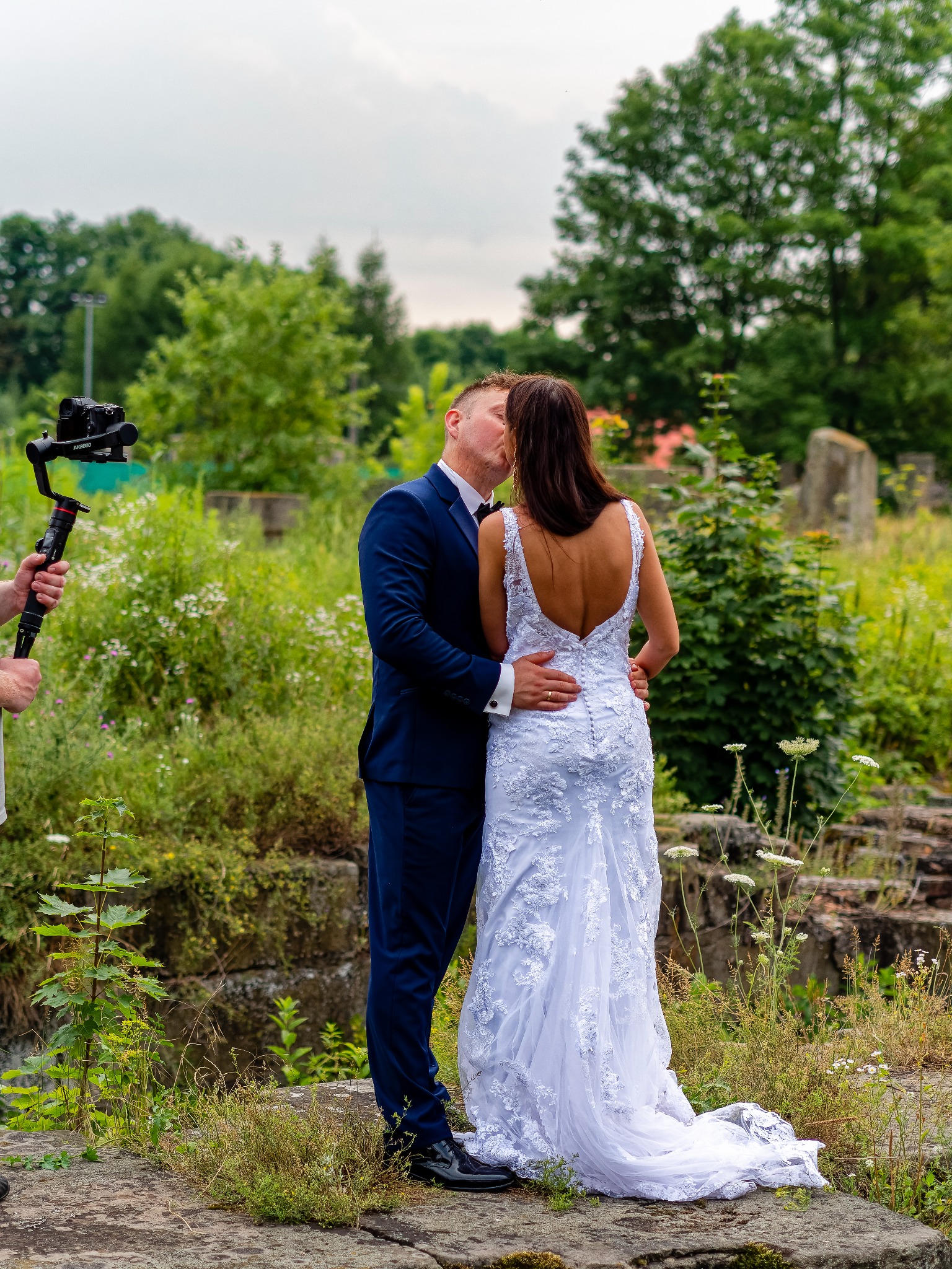 Pocałunek pary młodej w plenerze podczas filmowania z użyciem stabilizatora kamery, suknia ślubna z koronkowymi aplikacjami i głębokim dekoltem na plecach, granatowy garnitur pana młodego, zieleń...
