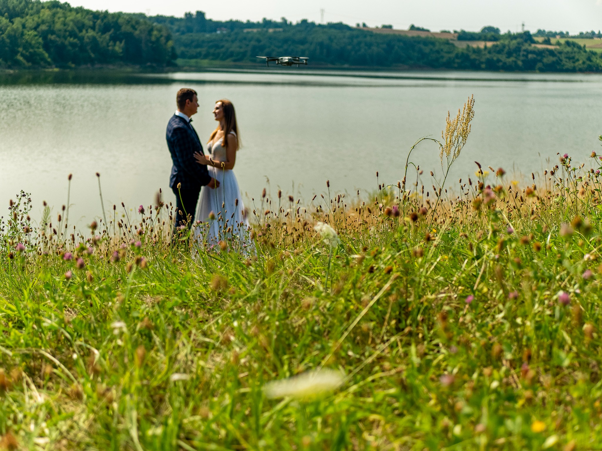 Para młoda w eleganckich strojach stoi na tle jeziora i zieleni, z dronem widocznym w tle, ujęcie z dołu przez wysokie trawy.