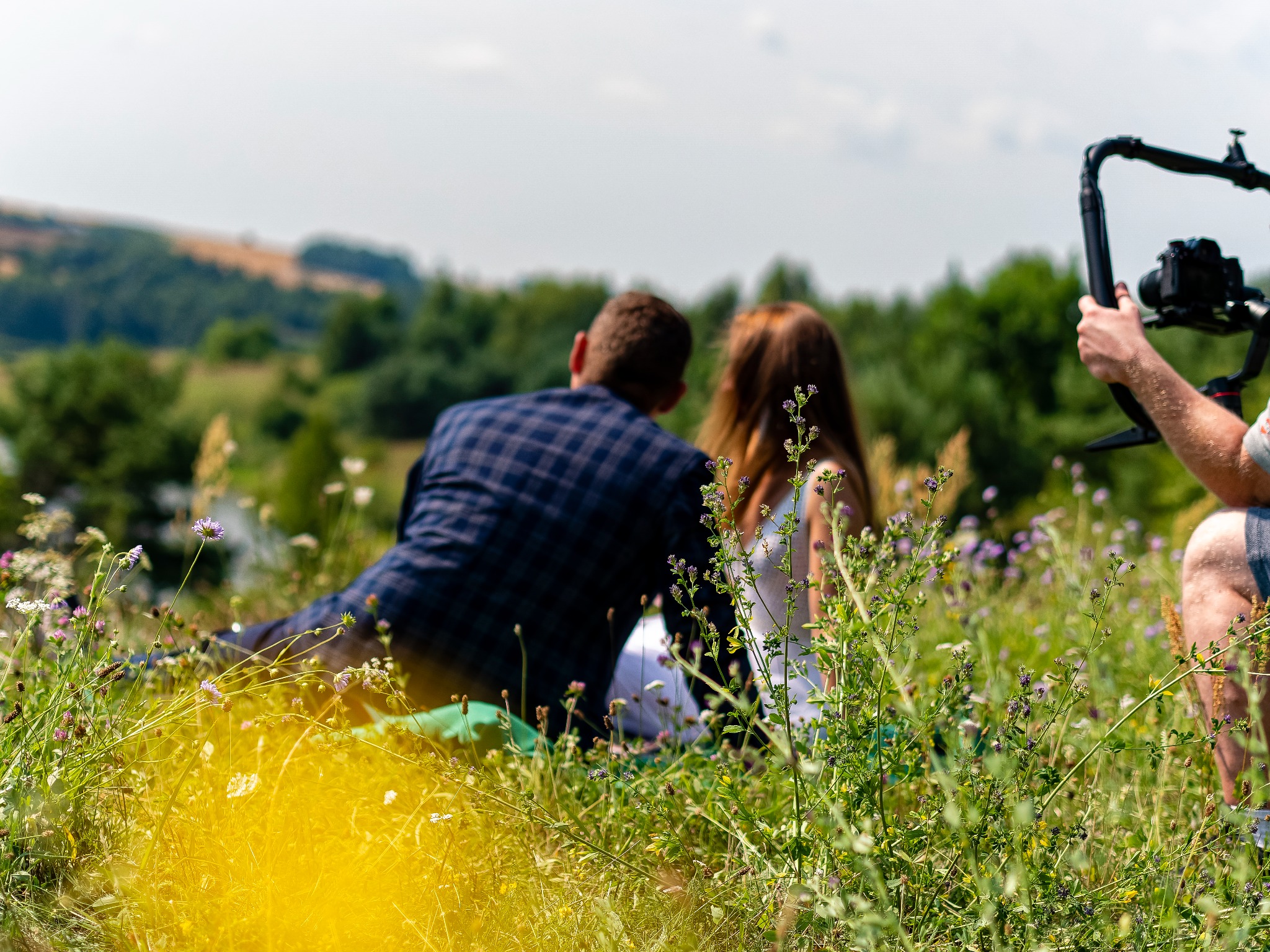 Para siedząca na łące podczas sesji zdjęciowej, widoczny operator kamery z rigiem, w tle zalesione wzgórza.